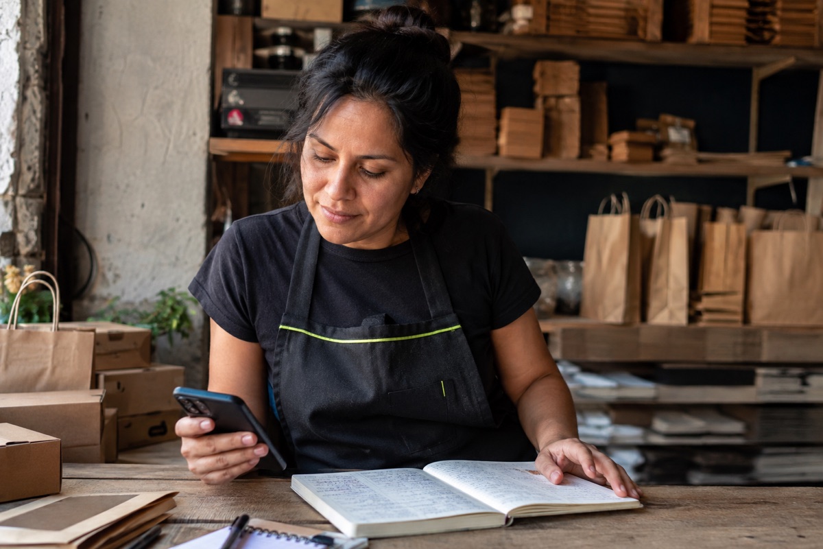 Escena ilustrativa: una comerciante revisa su teléfono y su libreta de pedidos.